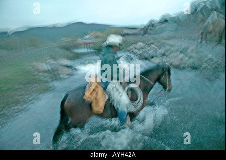 Am Ende von einem kalten regnerischen Tag kreuzt ein Cowboy einen Stream Überschrift zurück zur Ranch und zu Hause Stockfoto