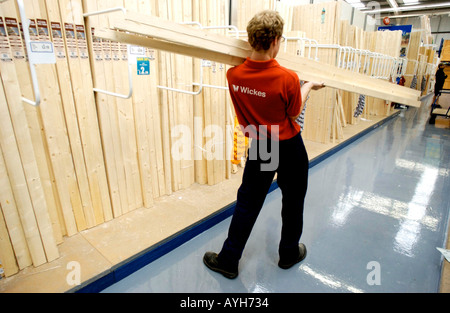 Junge männliche Assistant bei einem Baumarkt Wickes trägt ein Brett aus Holz Stockfoto