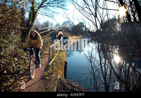 Lächelnde Frau in gelber Jacke Radfahren entlang eines Pfades Fluss im Osten Londons in geringer Sonneneinstrahlung an einem kalten Frühlingstag. Stockfoto