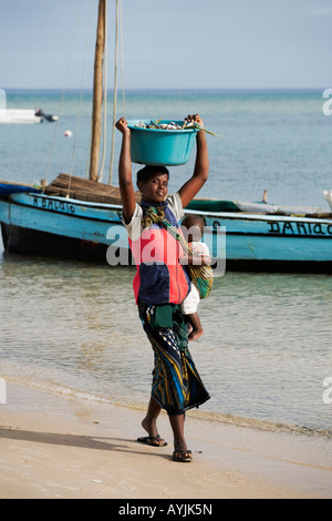 Afrikanische Frau mit Baby tragen Eimer gefüllt mit Krabben auf Kopf Dhow Segeln Boot im Hintergrund Benguerra Insel Mosambik Stockfoto