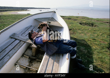 Bootsmann entspannt sich mit seinem kleinen Hund in einem Boot, Askam, Cumbria, England Stockfoto