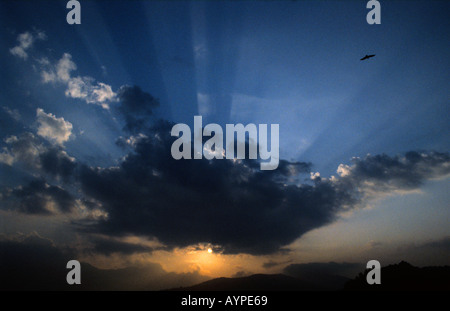 Malerischer Sonnenuntergang mit Rochen und fliegendem Vogel über den Silhouetten der Berge in der Abenddämmerung. Dramatische kammerköpuskuläre Strahlen durchdringen dunkle Wolken in einem blauen Himmel. Stockfoto