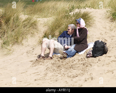 Älteres Ehepaar sitzt auf Sanddünen am Summerleaze Beach, Cornwall, an einem windigen Frühlingstag. Stockfoto