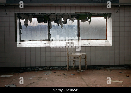 Schmutziges altes Fenster in einem verlassenen, gefliesten Zimmer mit einem abblätternden weißen Stuhl und Schutt auf dem Boden. Stockfoto