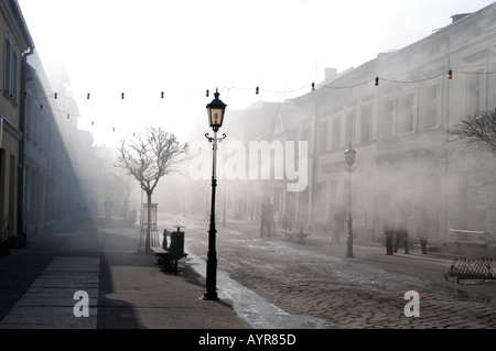 Atmosphärischer Nebel am Morgen auf der Szeroka Straße in Grodzisk Wielkopolski, Wielkopolska, Polen. Historische kopfsteingepflasterte Straße mit Vintage-Lampen. Stockfoto