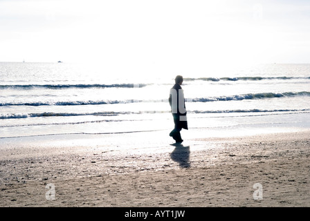 ein einsamer Mann im dunklen Mantel Spaziergänge an einem Strand in der Nähe von der Brandung in hellem Sonnenlicht Stockfoto