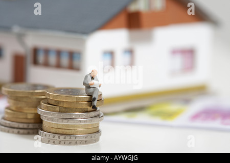 Geschäftsmann Figur sitzt auf Stapel Münzen während des Lesens, Miniaturhaus im Hintergrund Stockfoto