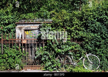 Ein altes Fahrrad sitzt vor rostigen Eisenzaun, bedeckt mit Reben, mit alten Zement Gebäude im Hintergrund Stockfoto