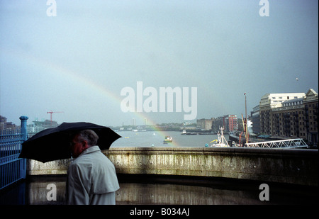 Ein Mann mit einem Regenschirm Spaziergänge während ein Sturm mit Regenbogen im Hintergrund, London, UK, 15.05.2000. Stockfoto