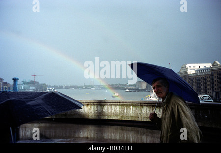 Ein Mann mit einem Regenschirm Spaziergänge während ein Sturm mit Regenbogen im Hintergrund, London, UK, 15.05.2000. Stockfoto