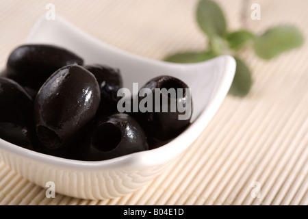 kleine weiße Schale mit schwarzen Oliven ohne Kern, auf Strohmatte, garniert mit Blättern Stockfoto