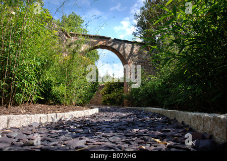 EIN STEIN BOGEN FÜHRT ZU EINEM GARTENWEG GELEGT MIT SCHIEFER SCHERBEN UND EINGEFASST MIT GRANIT PFLASTERSTEINE Stockfoto