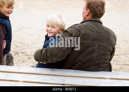 Ein liebender Vater sitzt mit seinen beiden Söhnen, während sie zusammen während einem Tag am Strand herumtollen Stockfoto