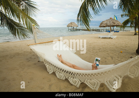 Frau in Hängematte zwischen Palmen am Strand mit strohgedeckten Palapas von Karibik, Jaguar Reef Lodge, Hopkins liest Stockfoto