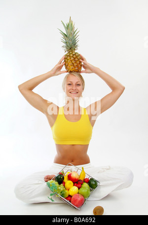 junge sportliche Frau in einem gelben Top mit Obstkorb und eine Ananas auf dem Kopf Stockfoto