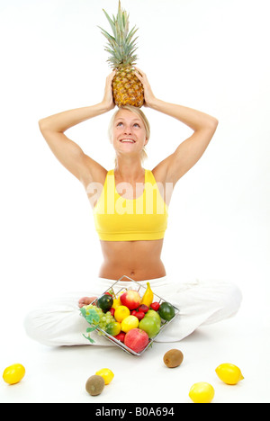 junge sportliche Frau in einem gelben Top mit Obstkorb und eine Ananas auf dem Kopf Stockfoto