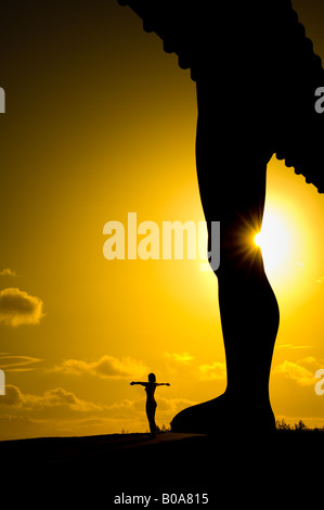 Silhouette einer Frau, die mit ausgestreckten Armen am Fuß des Engels des Nordens in Gateshead steht. Vor einem goldenen Himmel gesehen Stockfoto