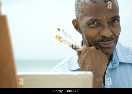 Ältere Mann mit Hand unter Kinn, hält Pinsel, lächelnd in die Kamera, Nahaufnahme Stockfoto