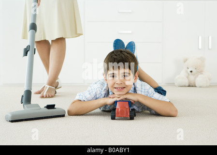 Kleiner Junge auf dem Boden liegend mit Spielzeug, lächelnd in die Kamera, Mutter saugen um ihn herum Stockfoto