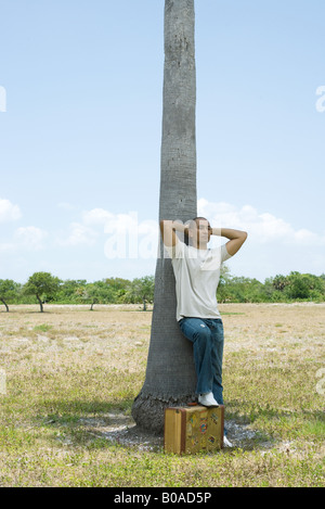 Mann gelehnt Baumstamm, Hände hinter dem Kopf, einen Fuß ruht auf Koffer Stockfoto