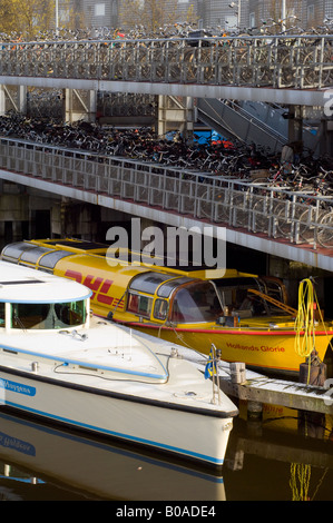 Amsterdam, Fahrräder geparkt in einem Fahrradpark am Stationsplein und zwei Boote vertäut am offenen Hafen Kanal Stockfoto