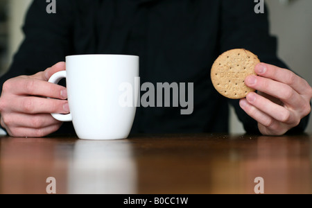 Eine Person sitzt an einem Küchentisch mit einer Tasse Tee und einen Keks. Stockfoto