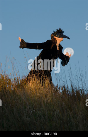 Eine maskierte Frau in einem schwarzen Samtkleid tarnt sich als eine Krähe oder Rabe Geist mit dem Vollmond im Hintergrund Stockfoto