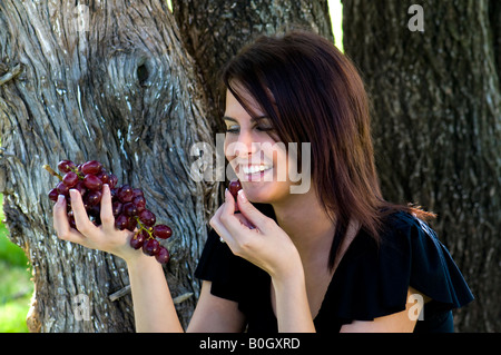 Eine schöne junge Frau sitzt unter Bäumen und Weintraube in der Hand hält, während der Vorbereitung zu einem Essen. Stockfoto