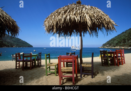 Bucht und Strand mit Holztischen und Stühlen unter reetgedeckten Sonnenschirmen, Bahia de Banderas, Playa Yelapa in der Nähe von Puerto Vallarta, J Stockfoto