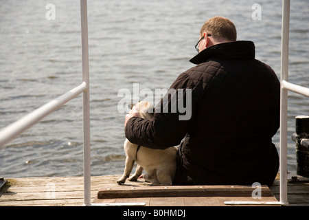 Mann und ein Mops sitzt auf einem hölzernen Dock, schaut sich um Stockfoto