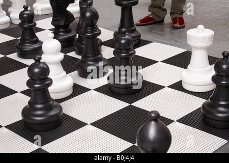 Blick auf einen übergroßen Schachbrett und Schach Stücke mit eines Spielers Füße entlang dem Rand des Brettes gesehen. Stockfoto