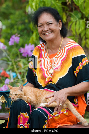 Eine bunt gekleideter älterer Thai Dorf Frau genießt sitzen in ihrem Garten mit Tabby Katze, Süd-Thailand. Stockfoto