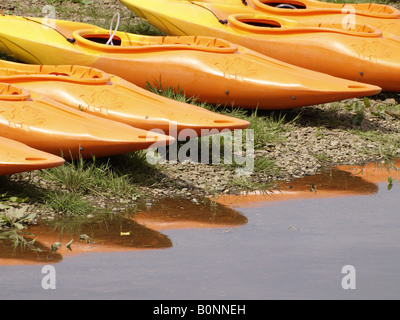 Reihe von orange Kajaks aufgereiht am Rande eines Flusses mit einer Reflexion in das ruhige Wasser. Stockfoto