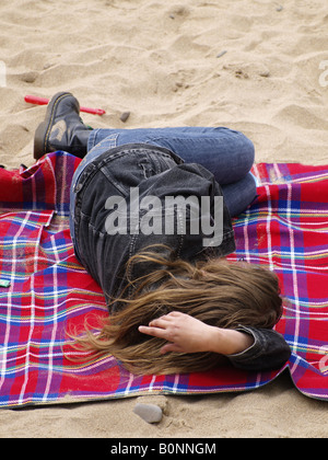 Junge Frau trägt Jeans, Jacke und Stiefel, Verlegung auf einer roten aufgegebenes Decke an einem Sandstrand in Cornwall Stockfoto