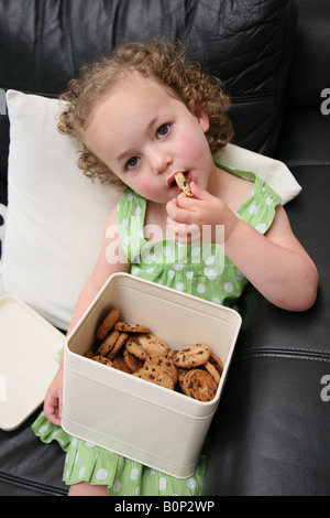 Ein 3 Jahres altes Mädchen auf dem Sofa zu Hause sitzen wird Gier und Eatings viele Kekse und Gebäck Stockfoto