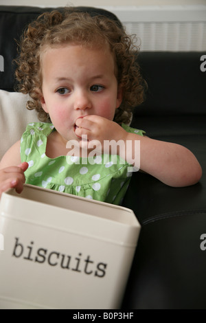 Ein 3-jähriges Mädchen sitzt auf dem Sofa zu Hause essen Cookies von einer Keksdose und overindulging Stockfoto