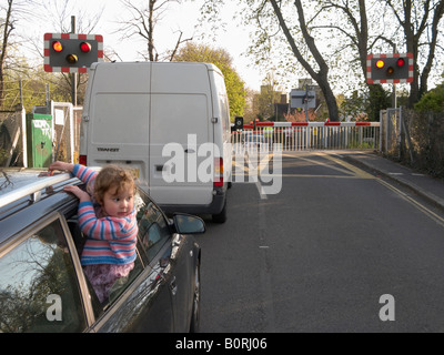 Fahrzeuge warten auf Eisenbahn Bahnübergang Barriere für Zug übergeben, & kleines Kind hängt gefährlich aus einem Autofenster zu beobachten. Stockfoto