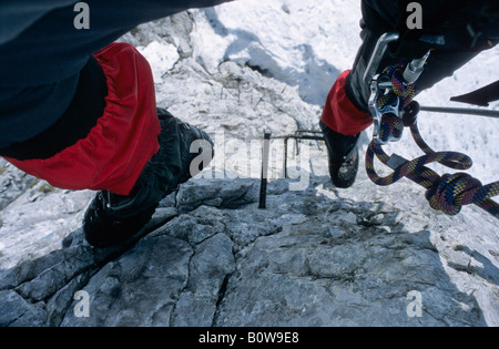 Seil und Karabiner, bedeckt Felsen klettern Satz sichtbar zwischen die roten Gamasche Beine von einem Kletterer Klettern auf Metall Sprossen Schinken Stockfoto