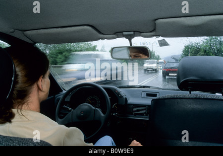 Frau sitzt auf einem Lenkrad fahren eine Auto, Regen und schweren Verkehr sichtbar durch Windschutzscheibe Stockfoto