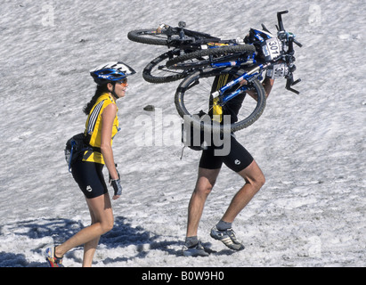 Mountain Bike Racer tragen zwei Mountainbikes durch den Schnee neben lächelnde weibliche Radfahrer tragen gelb, Adidas Bike Tran Stockfoto