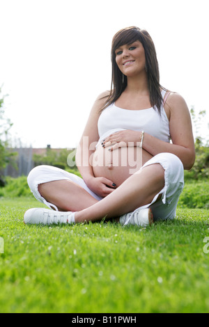 Happy-36-Woche, die schwangerer Teenager-Mädchen sitzen gekreuzt Beinen außerhalb der auf Rasen durch sich selbst und halten den exponierten Bauch Stockfoto