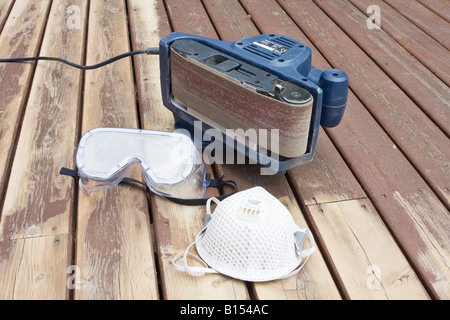 Bandschleifer auf seiner Seite mit abgenutzten Schleifband auf teilweise geschliffene Holzdeck bei Renovierung mit Schutzbrille und Maske Stockfoto