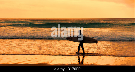 Ein Surfer seine board Silhouette gegen einen intensiven Sonnenuntergang, als er am Strand spazieren an Sennen in Cornwall. Stockfoto