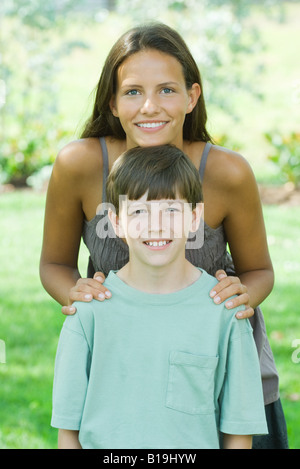 Teen Mädchen hinter dem jüngeren Bruder, ihr Kinn auf seinen Kopf, beide lächelnd in die Kamera ruht Stockfoto