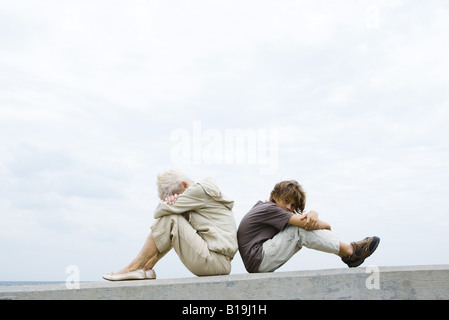 Großmutter und Enkel sitzen Rücken an Rücken, Kopf, Arme verschränkt, junge Blick in die Kamera Stockfoto