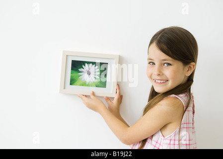 Kleines Mädchen hält gerahmtes Bild gegen die Wand, über die Schulter in die Kamera Lächeln Stockfoto