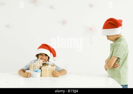 Boy holding Stack von Weihnachtsgeschenken, Bruder stehen in der Nähe, schmollen, beide tragen Santa Hüte Stockfoto