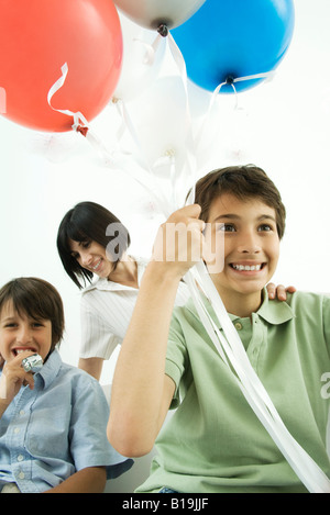 Junge mit Helium-Ballons, lächelnd, Mutter und Bruder im Hintergrund Stockfoto