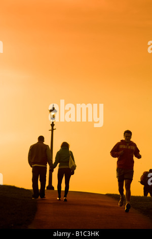 Paar gehen hand in hand, Primrose Hill bei Sonnenuntergang, während ein Jogger vorbei läuft Stockfoto