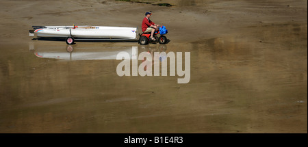 EIN MANN SCHLEPPT EIN GROßES KANU MIT EINEM KLEINEN MOTORRAD AUF EINEM WALISISCHEN STRAND, UK. Stockfoto